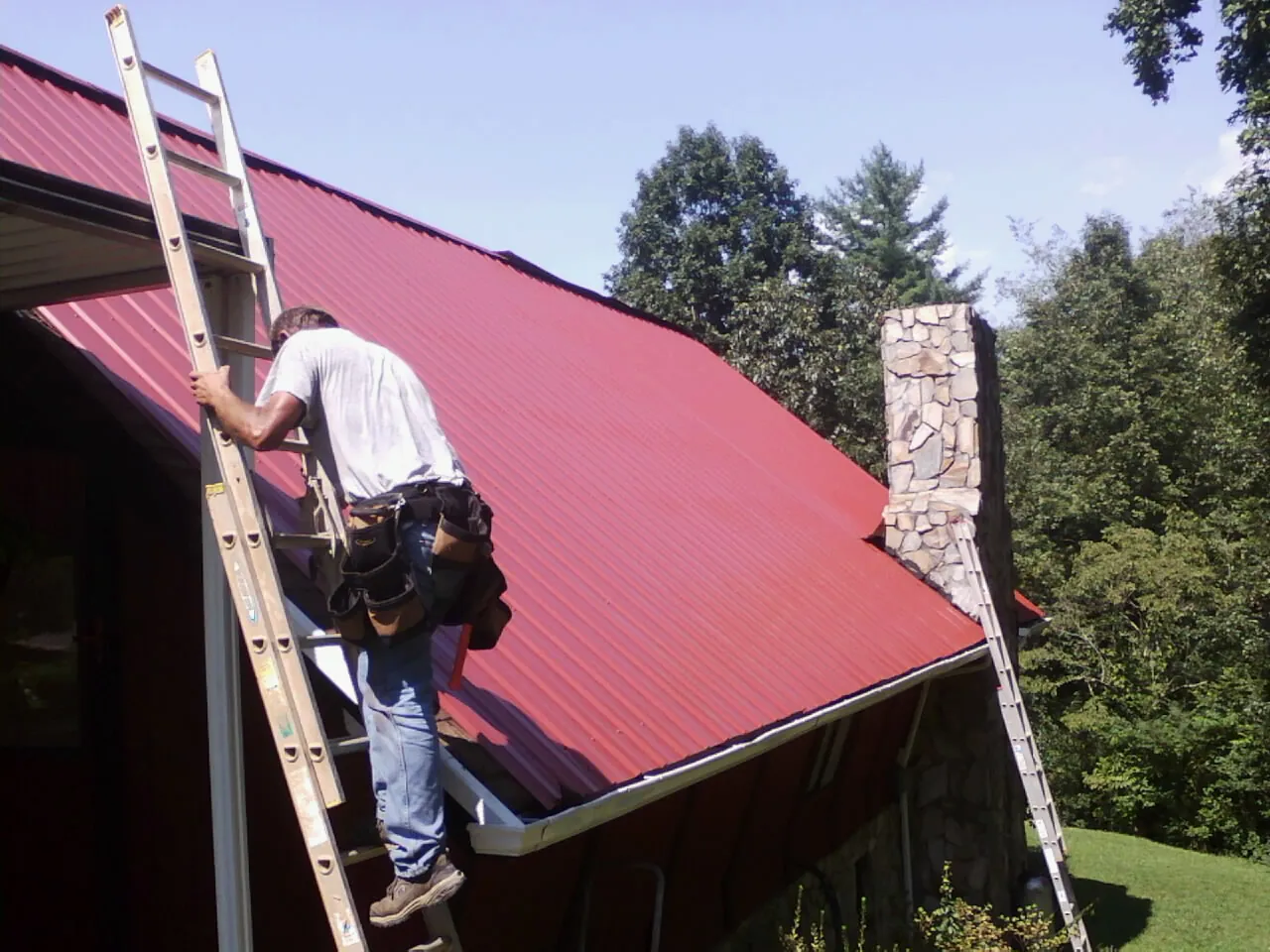 Roofer on a ladder beside a red metal roof and stone chimney — Soffit & Fascia Repair by Roofing Brothers in Smiley